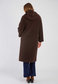Brązowy płaszcz polarowy o luźnym kroju i kapturze, z długimi rękawami i średnią długością. Noszony na granatowe szerokie spodnie.