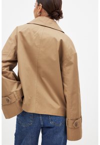 Bärnstenfärgad bomullsjacka med bred krage och oversized ärmar. Har stora manschettknappar och en slät textur. Kombineras med blå denimjeans.