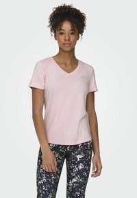Camiseta de manga corta en V de color rosa claro, hecha de una tela suave, con un pequeño detalle de logo en el pecho. Combinada con leggings de estampado floral.