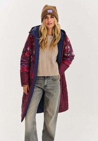 Pikowany długi płaszcz z kwiatowym wzorem w kolorach bordowym i granatowym, zestawiony z beżowym swetrem w splot warkocza i luźnymi szarymi jeansami. Brązowa czapka typu beanie.