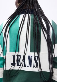 Άτομο με μακριές μαύρες κοτσίδες, φοράει πράσινο και άσπρο ριγέ πουκάμισο με τη λέξη "JEANS" στην πλάτη, κοιτάζοντας προς τα πίσω.