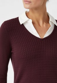 Frau, die einen tiefpurpurfarbenen strukturierten V-Ausschnitt-Pullover über einem weißen Hemd mit Kragen trägt, zu sehen von Hals bis zur Mitte des Oberkörpers.
