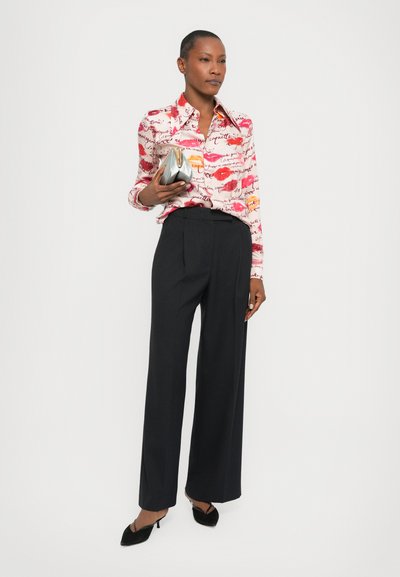 Long-sleeve, pink shirt with red lip prints and text, paired with black, wide-leg pinstripe trousers. Holds a silver clutch bag.