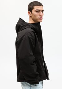 Schwarze wasserdichte Jacke mit Kapuze, mit einem Reißverschluss vorne, seitlichen Taschen und einer glatten, matten Textur. Lässiger Schnitt mit minimalistischer Gestaltung.