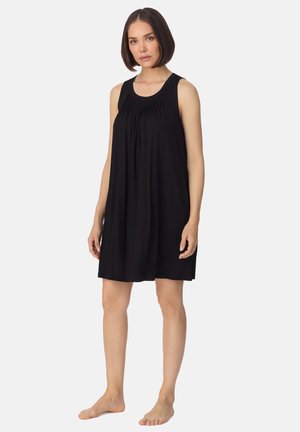 Vrouw staat blootsvoets en draagt een mouwloze, zwarte jurk tot op de knie met geplooide details op de borst voor een effen witte achtergrond.