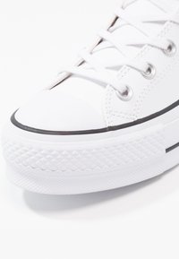 Gros plan sur une chaussure de sport basse blanche avec des lacets blancs, des œillets métalliques, une bande noire le long de la semelle en caoutchouc et un bout renforcé texturé.