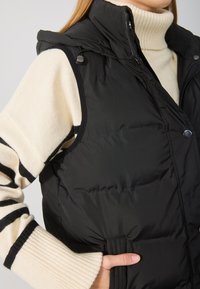 Schwarze gepolsterte Weste mit hohem Kragen und Druckknöpfen, getragen über einem creme- und schwarzgestreiften Pullover mit gerippten Bündchen.