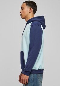 Világoskék és tengerészkék kapucnis sweatshirt, elülső zsebbel és bordázott mandzsettával. Puha anyagból készült, modern színblokkoló dizájnnal rendelkezik.