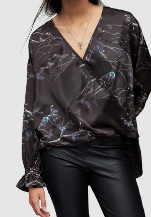 Femme portant un chemisier cache-cœur noir avec des motifs de papillons et de branches et un pantalon en cuir noir, visible des épaules jusqu'aux hanches.