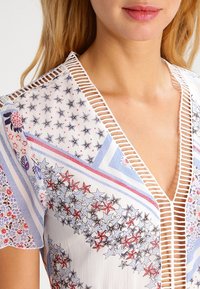 Blusa blanca con patrones de estrellas y flores multicolores, que presenta un profundo escote en V y elaborados detalles de corte a lo largo de los bordes.