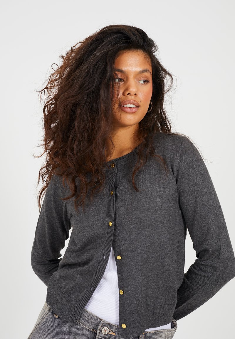 Gilet gris en tricot doux, avec un col rond, des manches longues et des boutons dorés sur le devant.