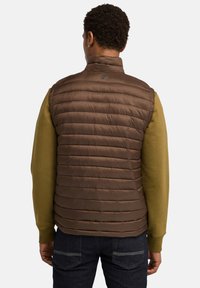 Bruine gevoerde puffer vest met hoge kraag, lichtgewicht materiaal en subtiele branding. Draagt over een groene lange mouwen shirt, gecombineerd met donkere spijkerbroek.