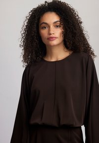 Pruun pikka varrukaga topp ümarate kaeluste jaoks, mille ees on pleat detail ja sile, voogav tekstuur.