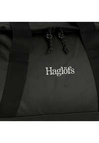 Zwarte sporttas met dubbele ritsen, stevig materiaal en brede webbing handgrepen. Voorzien van het geborduurde "Haglöfs" logo in zilver op de voorkant.