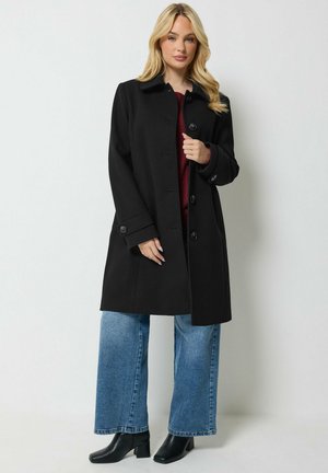 Femme aux longs cheveux blonds portant un manteau noir boutonné, un jean bleu à jambes larges, des bottes noires et un haut rouge sur un fond clair uni.