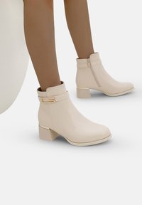 Botas de tobillo beige con un acabado suave, tacón cuadrado, acento dorado en la correa y cremallera lateral para un fácil calce.