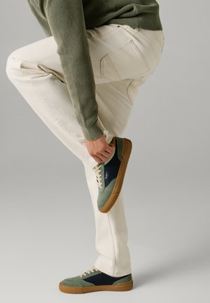 Oseba v kremnih hlačah in zeleni majici obuja zeleno-in-mornarsko supergo z rjavim podplatom na nevtralnem ozadju.