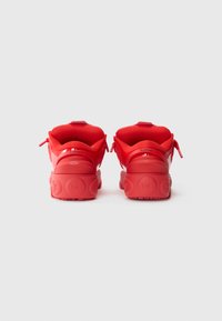 Un paio di scarpe da ginnastica rosse brillanti viste da dietro, con colli imbottiti e suole testurizzate con dettagli in rilievo, su uno sfondo bianco e semplice.