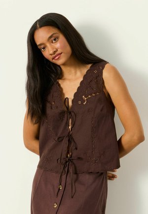 Junge Frau mit langen dunklen Haaren trägt ein ärmelloses braunes Oberteil mit Blumenstickerei und Bindeschließen, kombiniert mit einem passenden Rock mit Knöpfen.