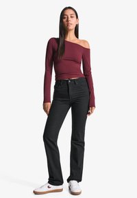 Burgundska bluzica brez naramnic z dolgimi rokavi, kombinirana z visokim pasom črnimi hlačami in belimi športnimi copati s svetlo rjavimi detajli na enobarvnem ozadju.