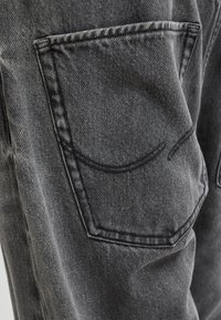 Czarne dżinsy z denimu z tylną kieszenią ozdobioną przeszyciami wzdłuż obrysu. Tkanina ma wyraźną fakturę, z widocznymi wzorami splotu.