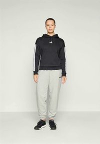 Sweatshirt preto com capuz e riscas brancas nas mangas, combinado com calças de treino cinzentas claras. Apresenta o logótipo à frente e ao lado. Sapatos atléticos pretos.