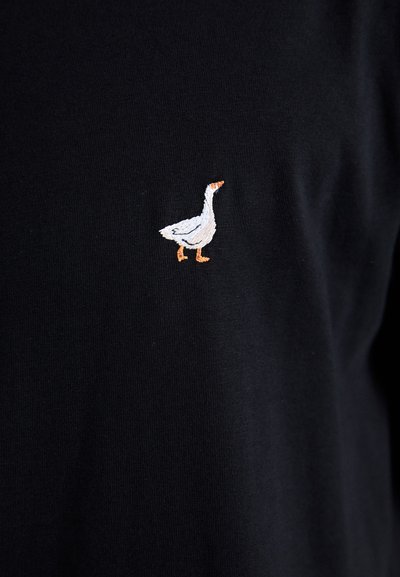 T-shirt en coton noir avec un petit oie blanche brodée sur la poitrine gauche, avec des détails sur le bec et les pattes en orange. Texture lisse.