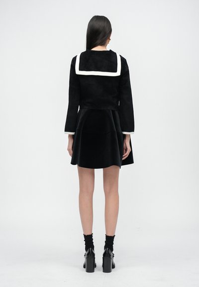 Schwarze Wollmischung Jacke mit weißem Matrosenkragen, kombiniert mit einem schwarzen Samt- Kleid mit ausgestellt Schnitt. Das Model trägt schwarze, hochhackige Stiefeletten und Socken.