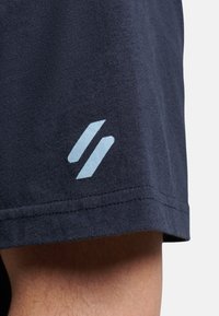 Pantaloni corti in cotone blu navy con un logo azzurro chiaro sull'orlo. Il tessuto ha una texture liscia con dettagli di cucitura puliti.