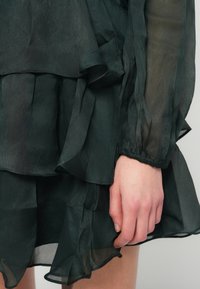 Γυναικεία χέρι ακουμπημένο πάνω σε στρωματοποιημένο σκούρο πράσινο διαφανές φόρεμα με ρυάκια στις άκρες και ελαστικά μανίκια.