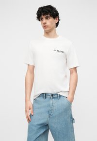 Νέος με σγουρά μαλλιά, ντυμένος με λευκό T-shirt Jack & Jones και ανοιχτό μπλε τζιν, στέκεται με το ένα χέρι στην τσέπη.