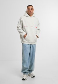 Světlé šedé oversized mikina s kapsou na přední straně a logovými detaily, v kombinaci s volnými světle modrými džínovými kalhotami a šedými teniskami.