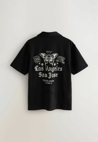 Mustat lyhythihaiset paita, jossa on hopeinen graafinen suunnittelu selässä, mukaan lukien perhonen, noppia ja teksti "Los Angeles San Jose Tattoo Studio."