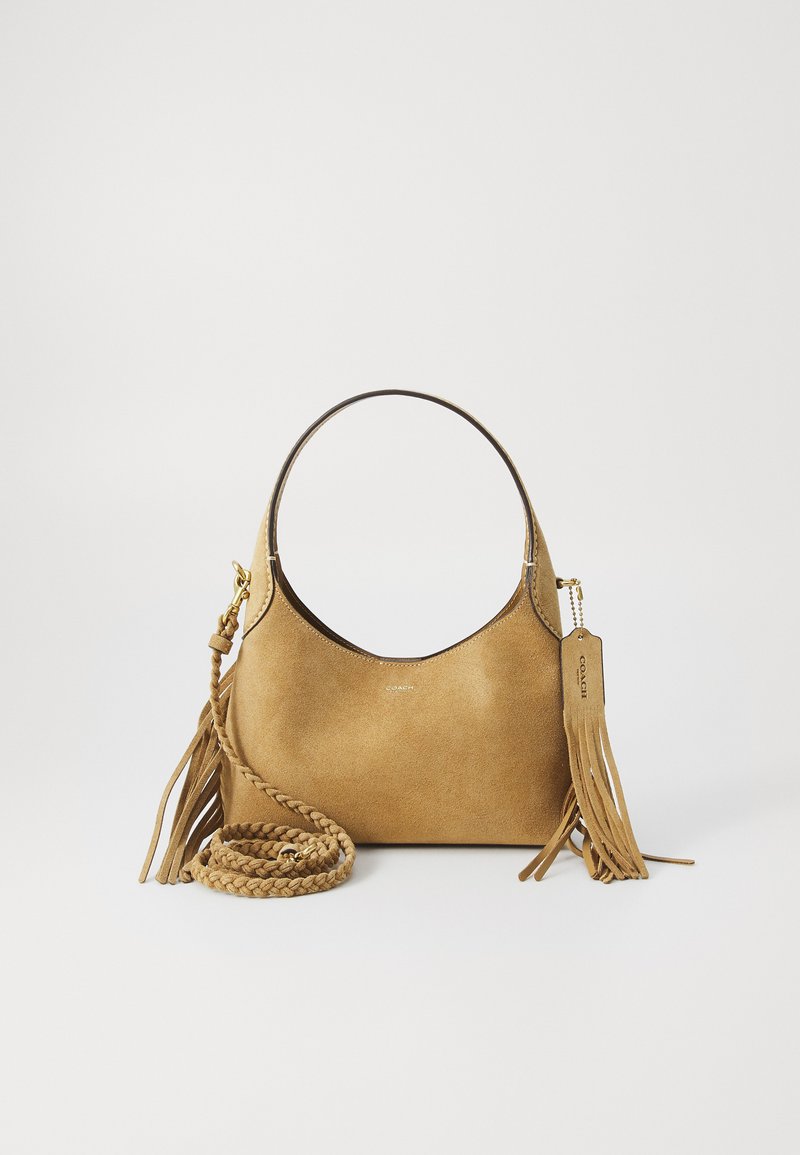 Sac à main en suede beige avec une forme arrondie, une bandoulière tressée et des franges décoratives. Comprend une étiquette logo et une texture lisse et douce.