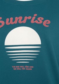 Türkis puuvillane T-särk, millel on valge päike ja kihilised jooned, koos sõnaga "Sunrise" roosana ning selle all inspireeriv tekst.