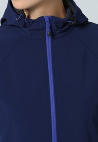 Chaqueta azul marino con capucha, que presenta una cremallera de longitud completa en contraste en color púrpura y un tejido suave. Detalles de costura a lo largo de los hombros.