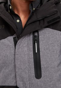 Černá a šedá bunda s texturovanou šedou látkou a pevným černým zipem. Obsahuje kapuci a viditelné logo v levém horním rohu.