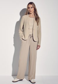 Beige blazer met een gestructureerde afwerking, gestreepte crèmekleurige en beige top, wijdvallende beige broek en witte sneakers met bruine zolen.