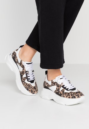 Personne portant des baskets blanches épaisses avec un imprimé léopard marron et noir, associées à un pantalon noir raccourci sur un fond uni.