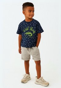 Niño de pie con las manos en los bolsillos, vistiendo una camiseta marina "Wild Thing", pantalones cortos gris claro y zapatillas beige, frente a un fondo blanco.