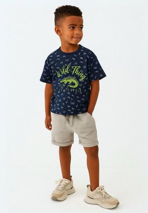 Pojke står med händerna i fickorna, iklädd en marinblå "Wild Thing" t-shirt, ljusgrå shorts och beige sneakers, mot vit bakgrund.