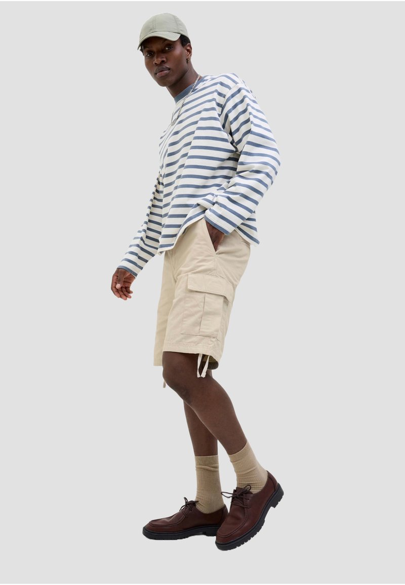Homme portant une casquette beige, une chemise à manches longues rayée, un short cargo beige, des chaussettes beiges et des chaussures marron, debout devant un fond uni.