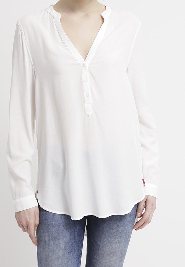 Blouse blanche à manches longues avec col en V et trois boutons, portée non rentrée sur un jean bleu par une personne debout.