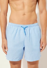 Světle modré pruhované plavky s elastickým pasem a šňůrkou na stažení. Vyrobeny z hladkého materiálu, mají boční kapsy.