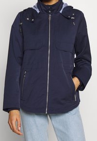 Marineblaue Jacke mit Reißverschluss vorne, zwei Brusttaschen und verstellbarem Kapuze. Gefütterte Kapuze mit einem blau-weißen Streifenmuster.