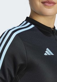 Schwarzes Sportoberteil mit hellblauen Streifen auf den Schultern, hohem Kragen und Reißverschluss. Hat ein kleines Logo auf der Brust.