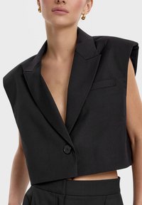 Czarna skórzana marynarka o krótkim kroju z głębokim dekoltem w kształcie V, poduszkami na ramionach, guzikiem na pojedyncze zapięcie oraz małą kieszonką na piersi. Materiał wydaje się gładki i strukturalny.