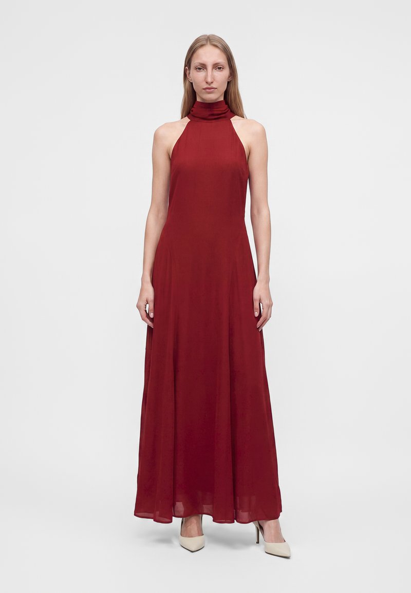 Femme debout portant une robe longue sans manches à col montant de couleur rouge foncé et des escarpins pointus crème sur un fond clair uni.