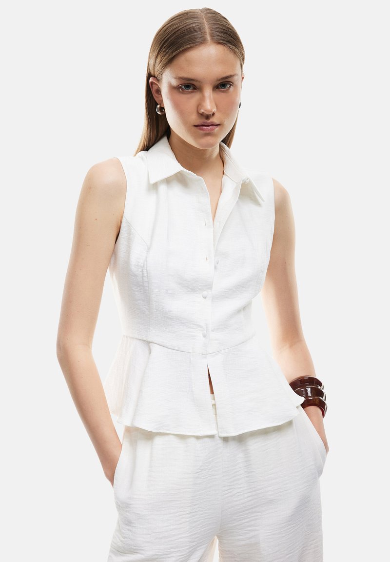 Hvid ærmeløs peplum top med knapfront, tekstureret stof, spids krave og en synlig underfold. Modellen bærer brune armbånd.