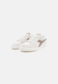 Beige suede sneakers with white rubber soles, featuring a brown leather side stripe and tonal laces. Includes a branded tan tongue tag.